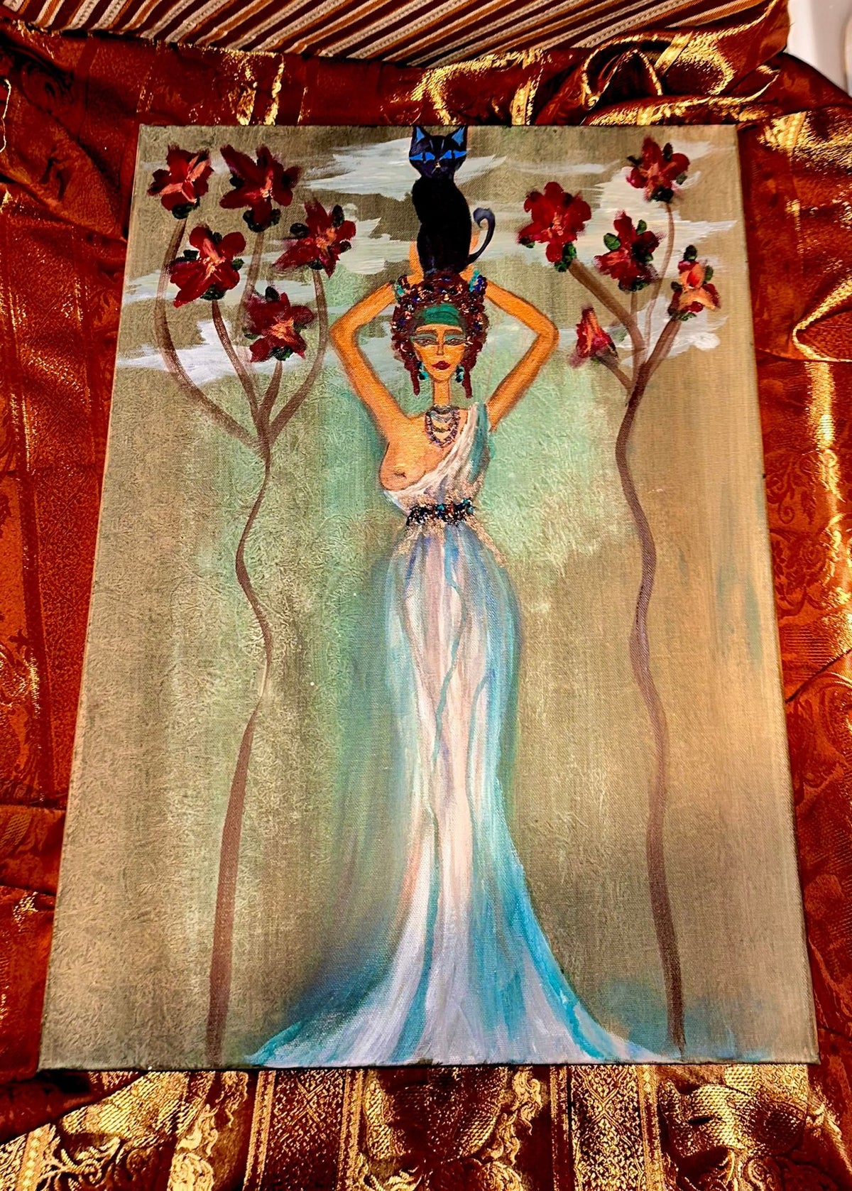 An Egyptian-style painting on canvas depicting a woman in a flowing white and blue gown holding a black cat on her head, surrounded by red flowers and gold-toned trees, displayed on a gold patterned fabric background.